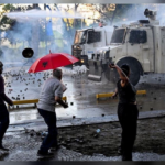 Riot police water cannon spraying protesters in Tirana, Albania, one holding a red umbrella with the Albanian double-headed eagle, SNN News Finland